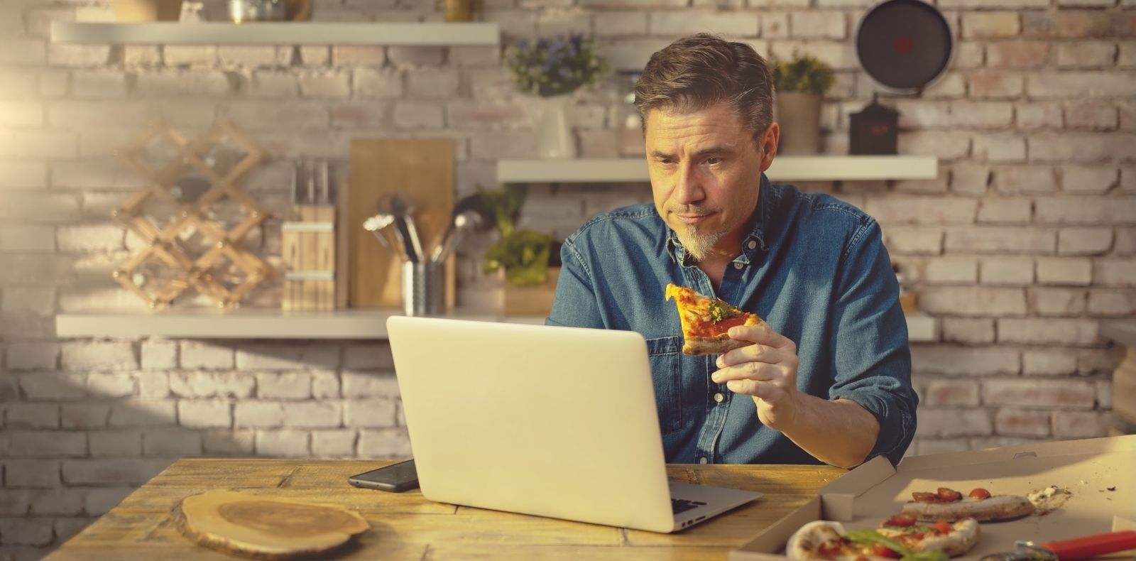 a man eating a slice of pizza while viewing his laptop